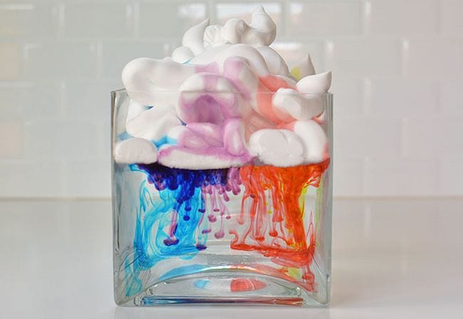 Square glass vase filled with water with shaving cream floating on top. Food coloring is dripping down through the cream into the water.